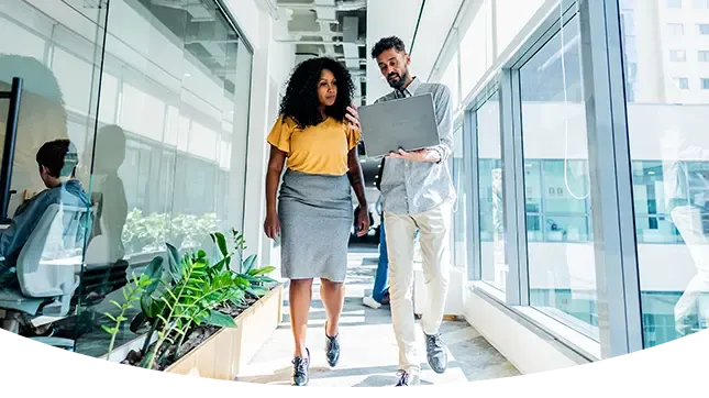 A man and woman walking down a bright, modern office hallway while looking at a laptop and discussing work.